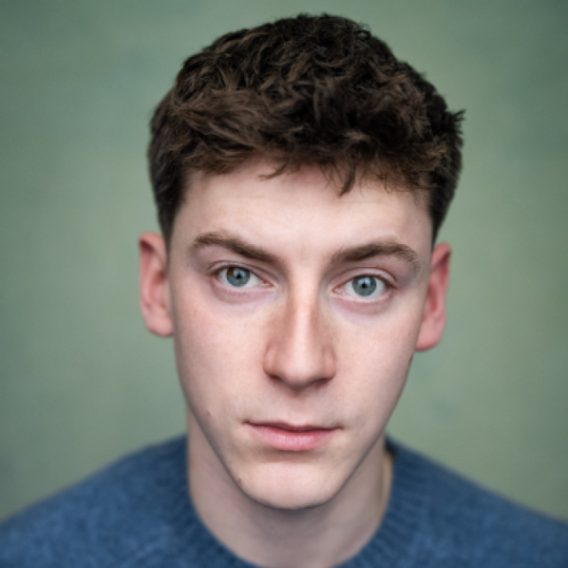 Close-up portrait of a young man with curly brown hair and blue eyes, wearing a blue jumper