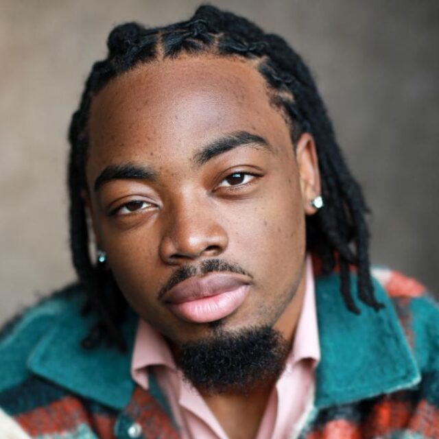 Close-up portrait of a man with shoulder-length locs and a short beard, wearing a teal jacket over a pink shirt
