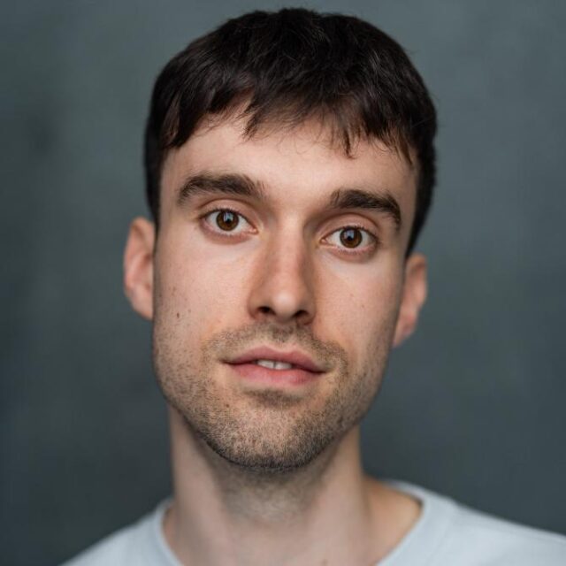 Close-up portrait of a young man with short dark hair and stubble, facing the camera against a grey background