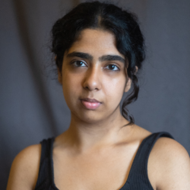 Portrait of a young woman with dark curly hair, wearing a black vest top, against a grey background