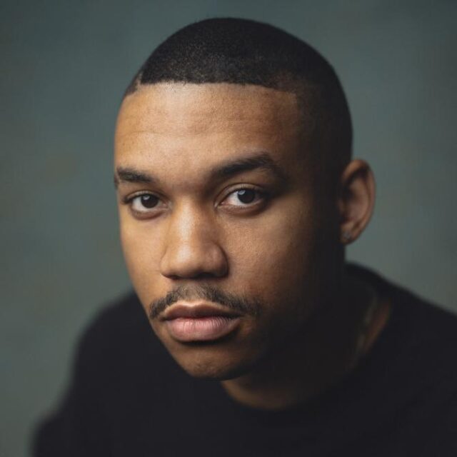 Close-up portrait of a young man with a short fade haircut, wearing a black top, looking directly at the camera with a neutral expression