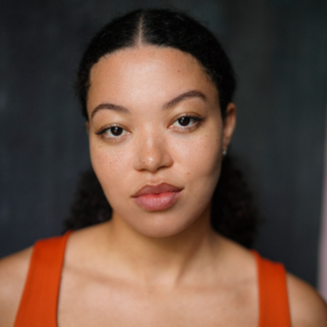 Close-up portrait of a woman with dark curly hair, wearing an orange top, against a dark background