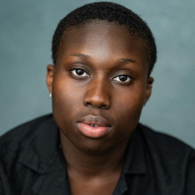 Close-up portrait of a person with short natural hair, wearing a dark shirt, looking directly at the camera