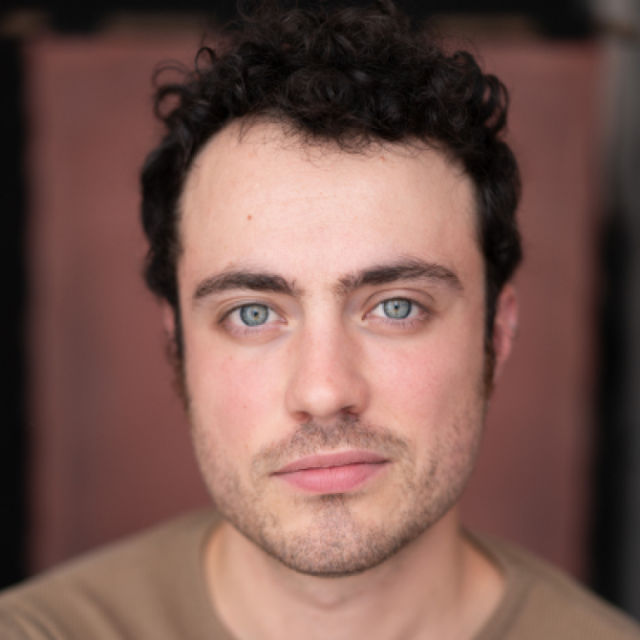 Close-up portrait of a young man with curly dark hair, blue eyes and light stubble, looking directly at the camera