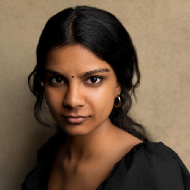 Close-up portrait of a young woman with long dark hair, wearing a black top and gold hoop earring, looking directly at the camera