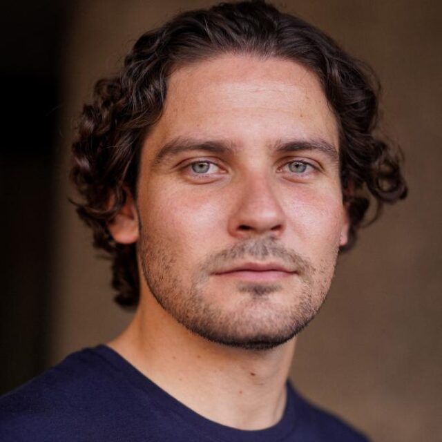 Man with curly dark hair and light stubble, looking directly at the camera