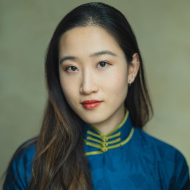 Portrait of a woman with long dark hair, wearing a blue traditional-style collared garment with gold frog fastenings