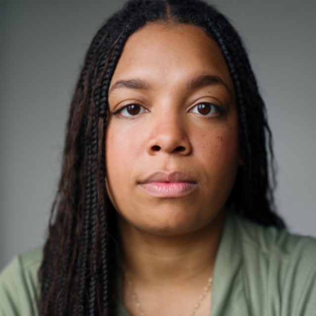 Close-up portrait of a woman with long braided hair, wearing a green top, looking directly at the camera with a neutral expression