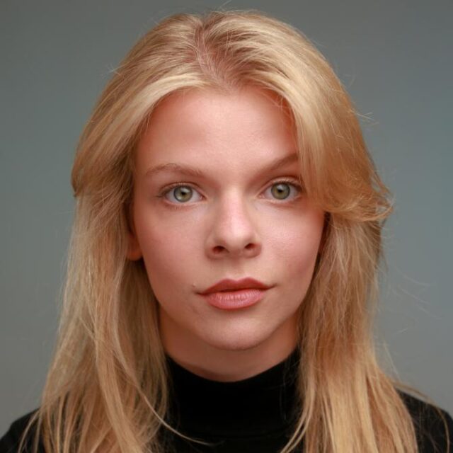 Portrait of a young woman with long blonde hair, wearing a black polo neck jumper, looking directly at the camera