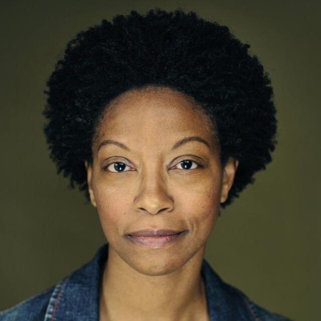 Close-up portrait of a woman with a natural afro, wearing a denim jacket, looking directly at the camera