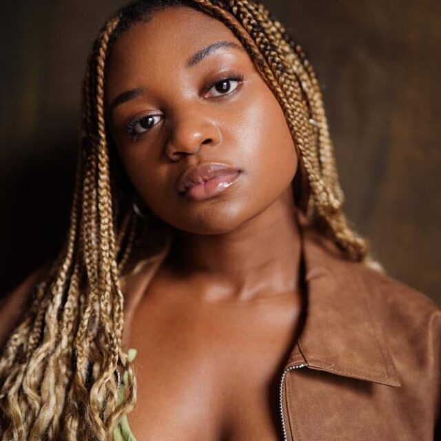 Woman with long blonde braids wearing a brown suede jacket, looking directly at the camera
