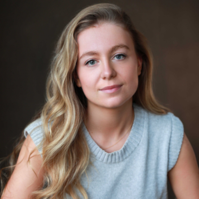 Portrait of a young woman with long wavy blonde hair, wearing a light grey sleeveless jumper