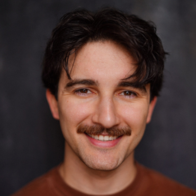Portrait of a smiling man with dark curly hair and a moustache, wearing a rust-coloured top