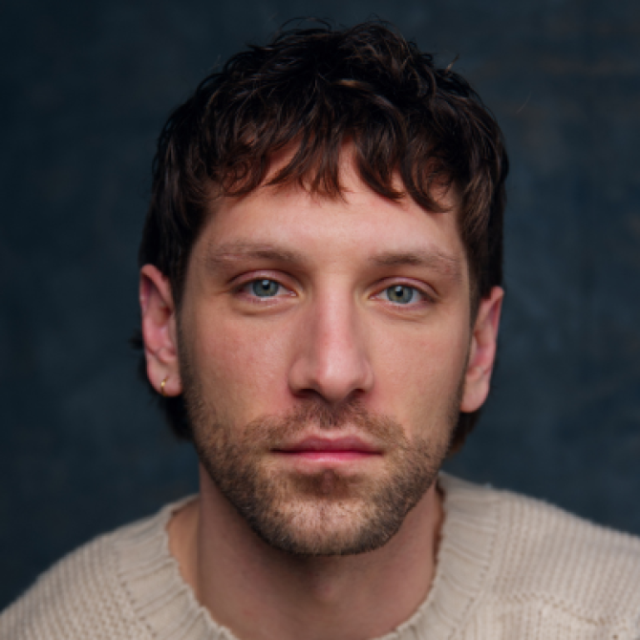 Man with curly dark hair and stubble, wearing a cream jumper, looking directly at the camera