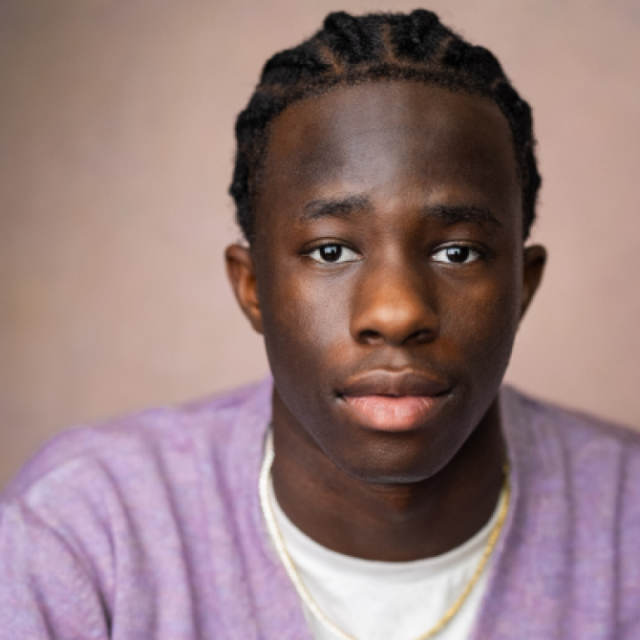 Close-up portrait of a young man with short braided hair, wearing a lilac jumper, looking directly at the camera with a neutral expression