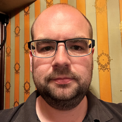 Close-up portrait of a bald man with glasses and a short beard, wearing a dark polo shirt, in front of a striped wallpaper