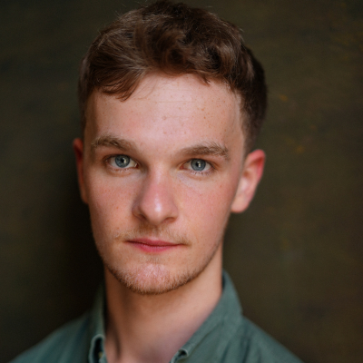 Headshot of a young man with wavy brown hair and light stubble, wearing a green shirt