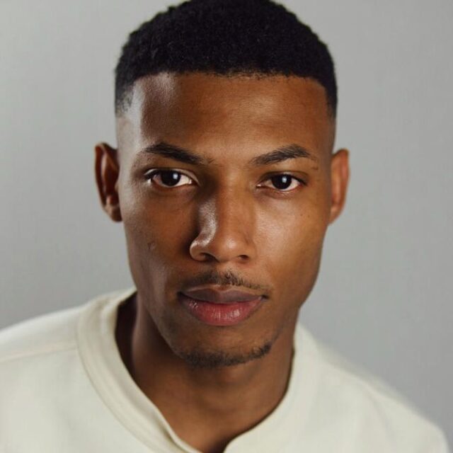 Close-up portrait of a man with short black hair, wearing a cream sweatshirt, looking directly at the camera