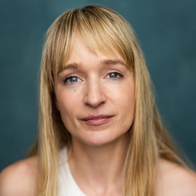 Close-up portrait of a woman with long blonde hair and a fringe, looking directly at the camera against a teal background