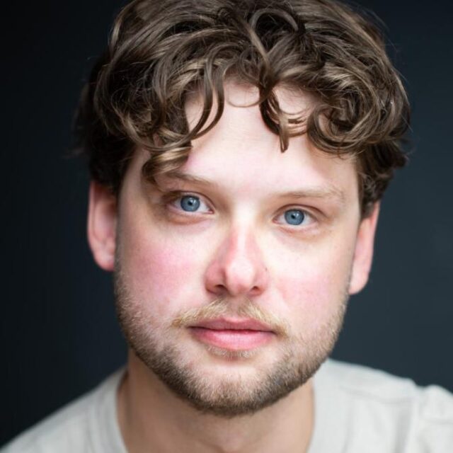 Close-up portrait of a young man with curly brown hair, blue eyes and light stubble, wearing a grey top
