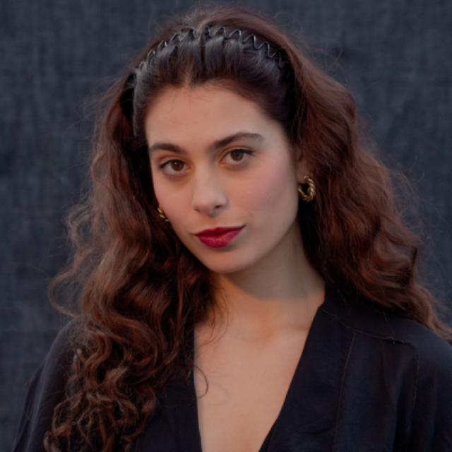 Woman with long curly dark hair, red lipstick and gold hoop earrings, wearing a black top