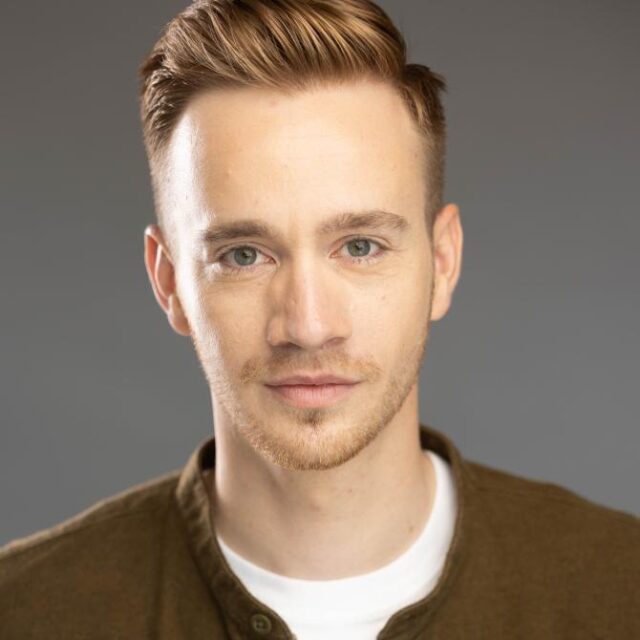 Headshot of a man with red hair and a beard, wearing a brown jacket over a white top