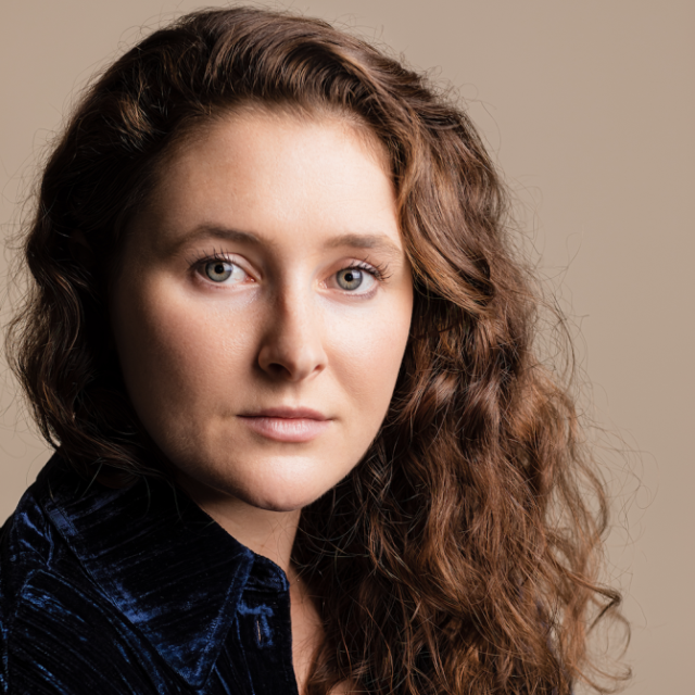 Portrait of a woman with long curly brown hair, wearing a dark blue velvet top, against a plain beige background