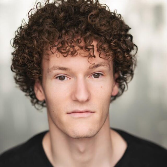 Close-up portrait of a young man with curly brown hair, wearing a black top, looking directly at the camera