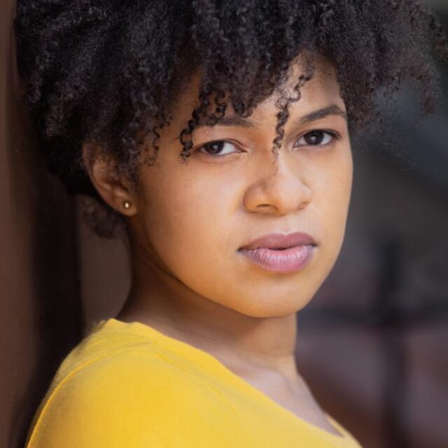 Close-up portrait of a woman with curly hair wearing a yellow top, looking directly at the camera with a serious expression