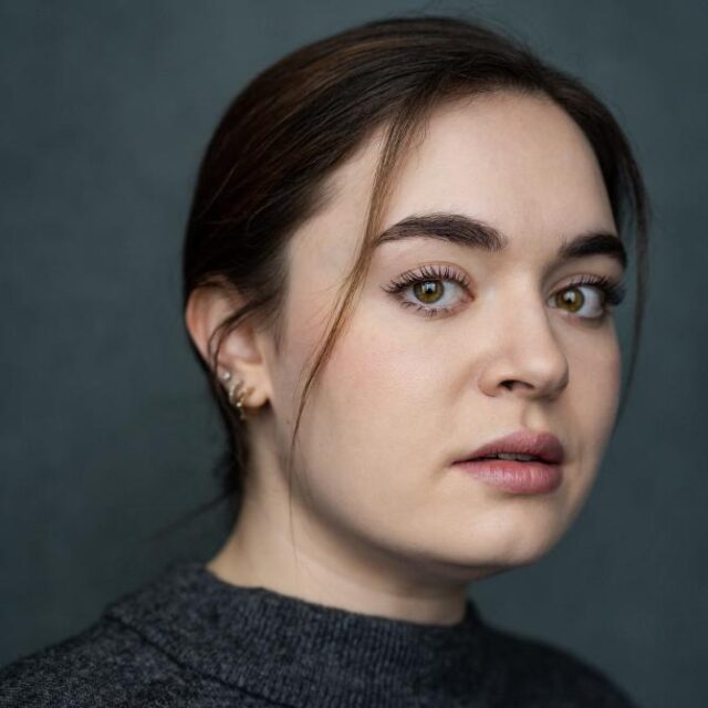 Close-up portrait of a young woman with dark hair, looking directly at the camera against a grey background