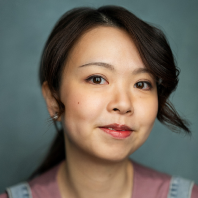 Close-up portrait of a woman with dark hair, smiling gently against a plain grey-blue background