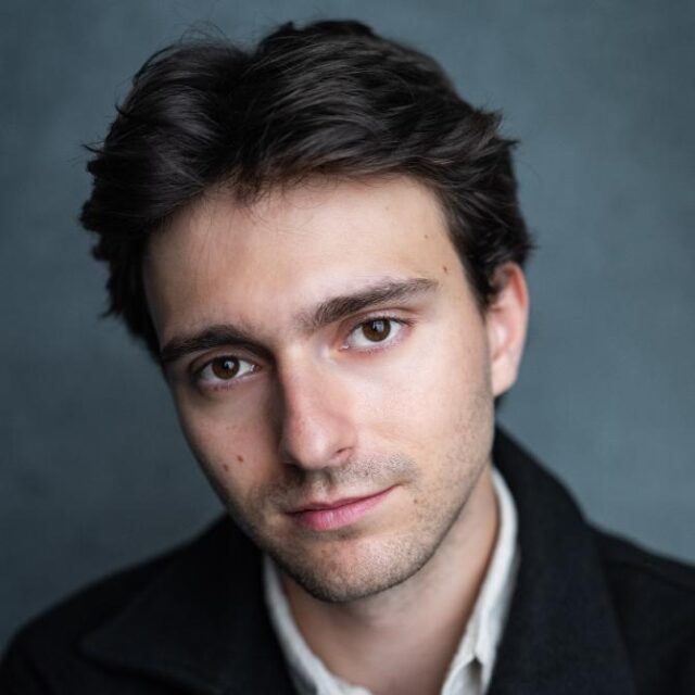 Close-up portrait of a young man with dark wavy hair, looking directly at the camera against a grey background
