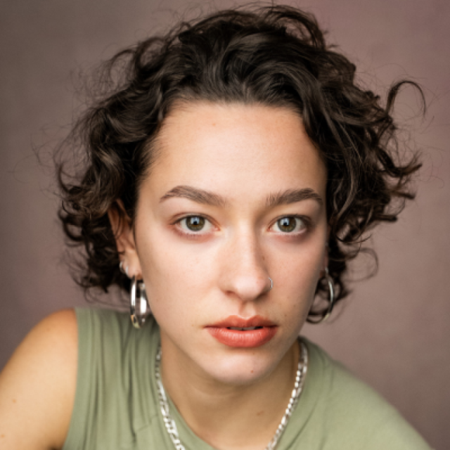 Woman with curly brown hair, hoop earrings and a chain necklace, looking directly at the camera