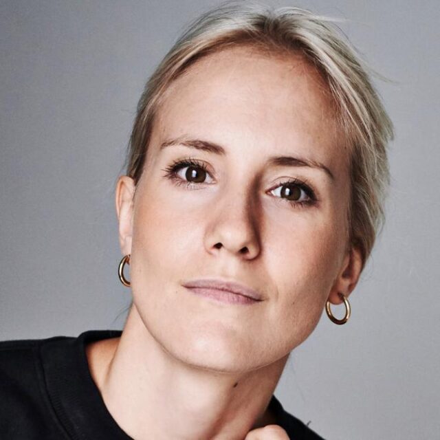 Close-up portrait of a woman with blonde hair and gold hoop earrings, looking directly at the camera