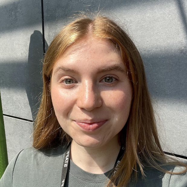 Close-up portrait of a young woman with long reddish-brown hair, smiling, wearing a lanyard outdoors