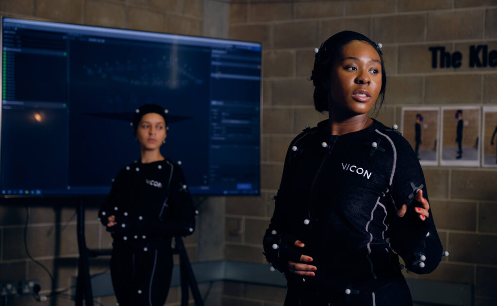 Two students in black motion capture suits stand in front of a large TV screen