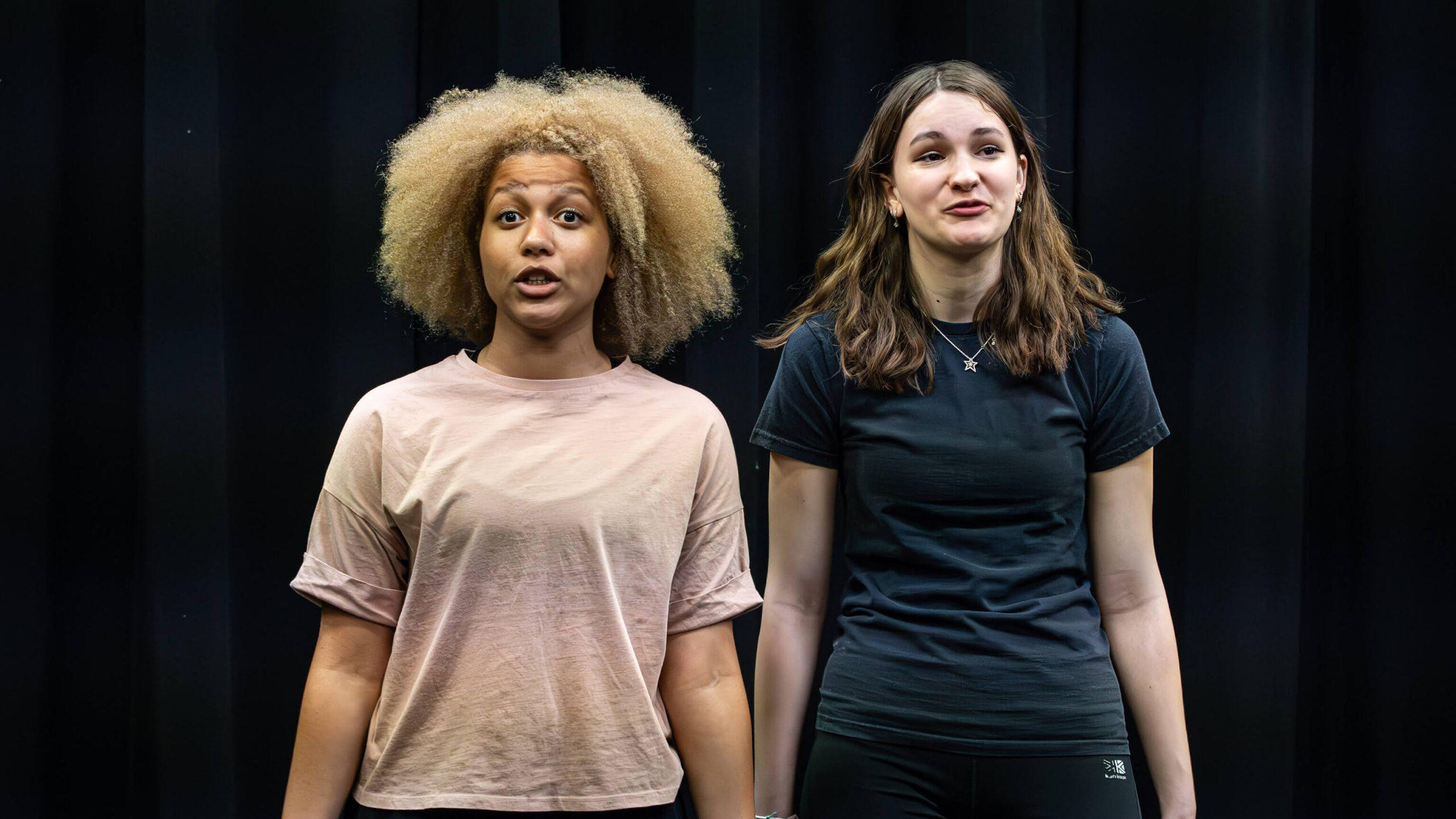 Two young women stand side by side on a stage against a black curtain, mid-speech during a performance or rehearsal